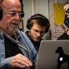 A professor with headphones works on a computer while two students also wearing headphones work alongside him.